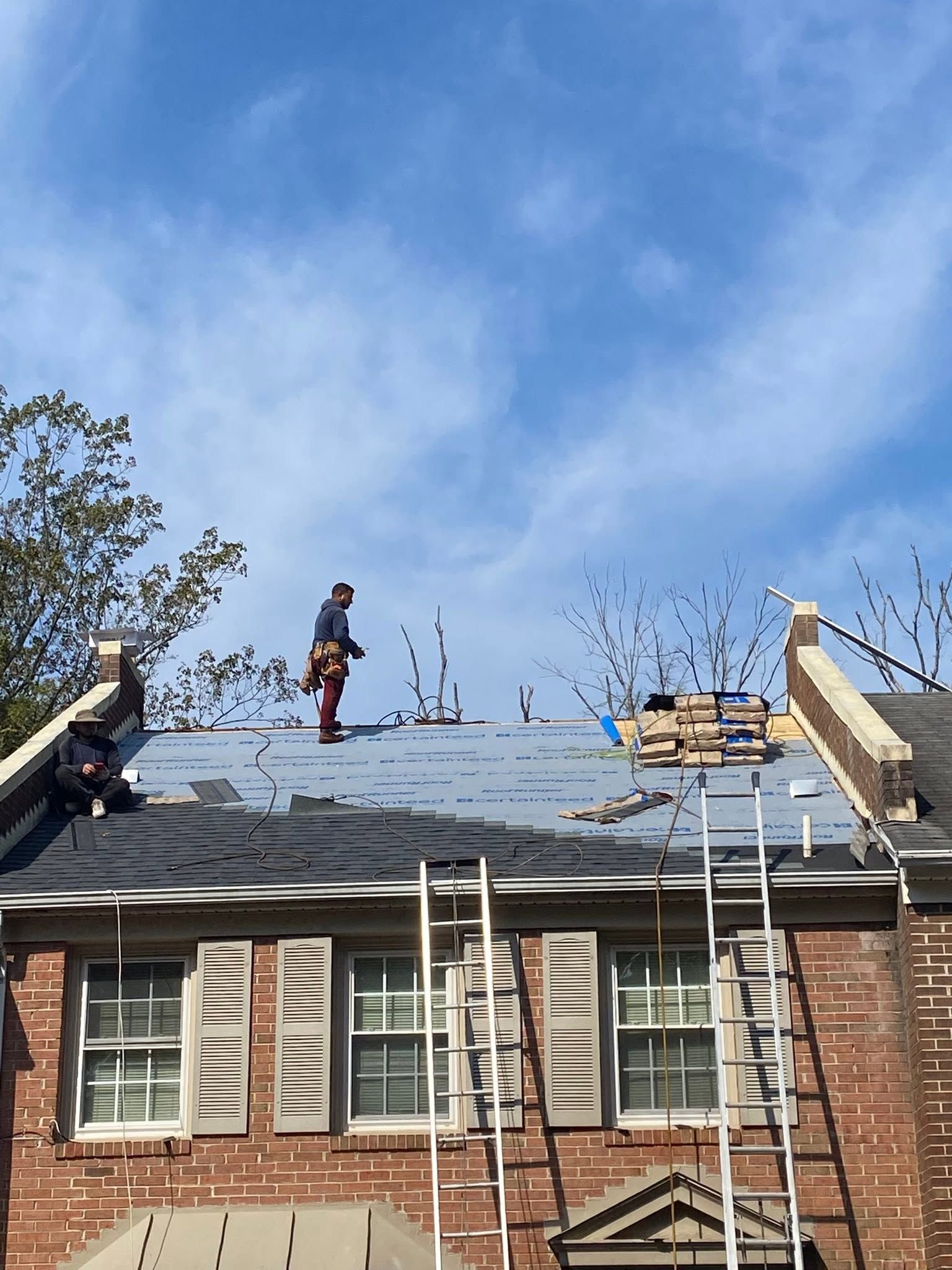 Roofers working on a brick house roof under a blue sky, with ladders and construction materials visible