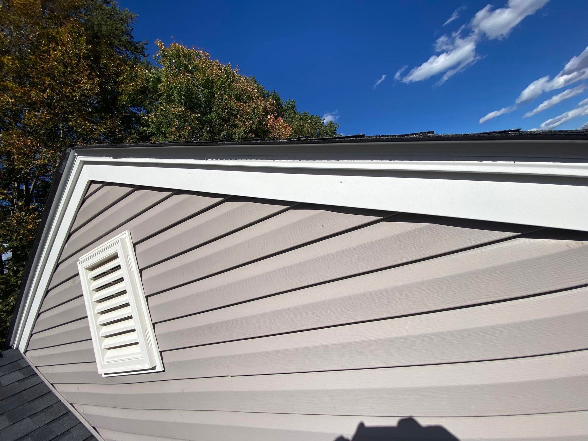White house siding with a vent under a blue sky and trees, viewed at an angle