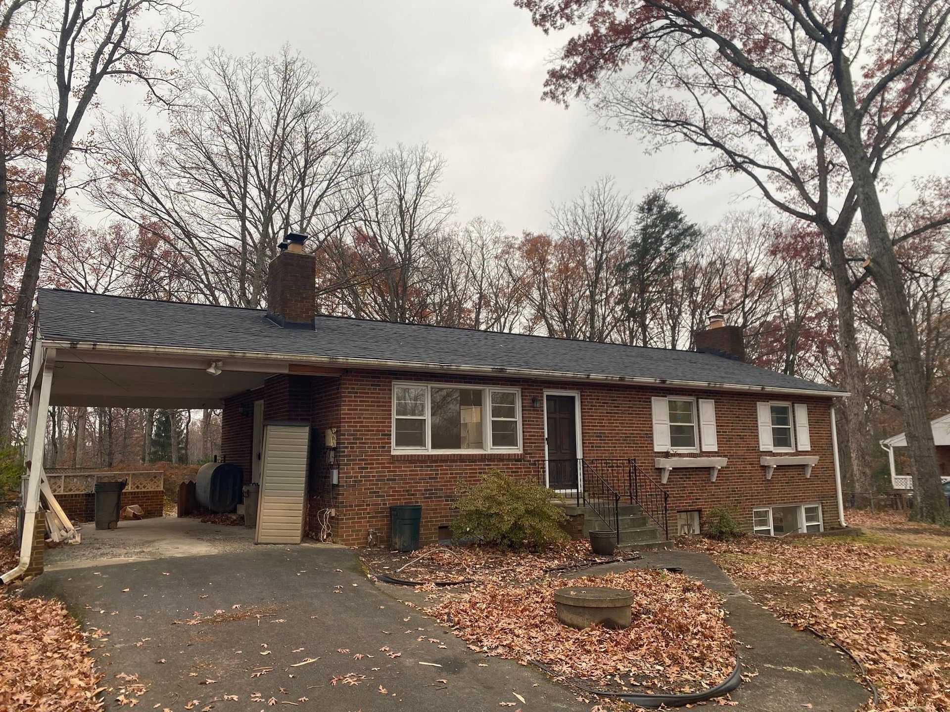 Single-story brick house with a carport, leaf-covered yard, and autumn trees.