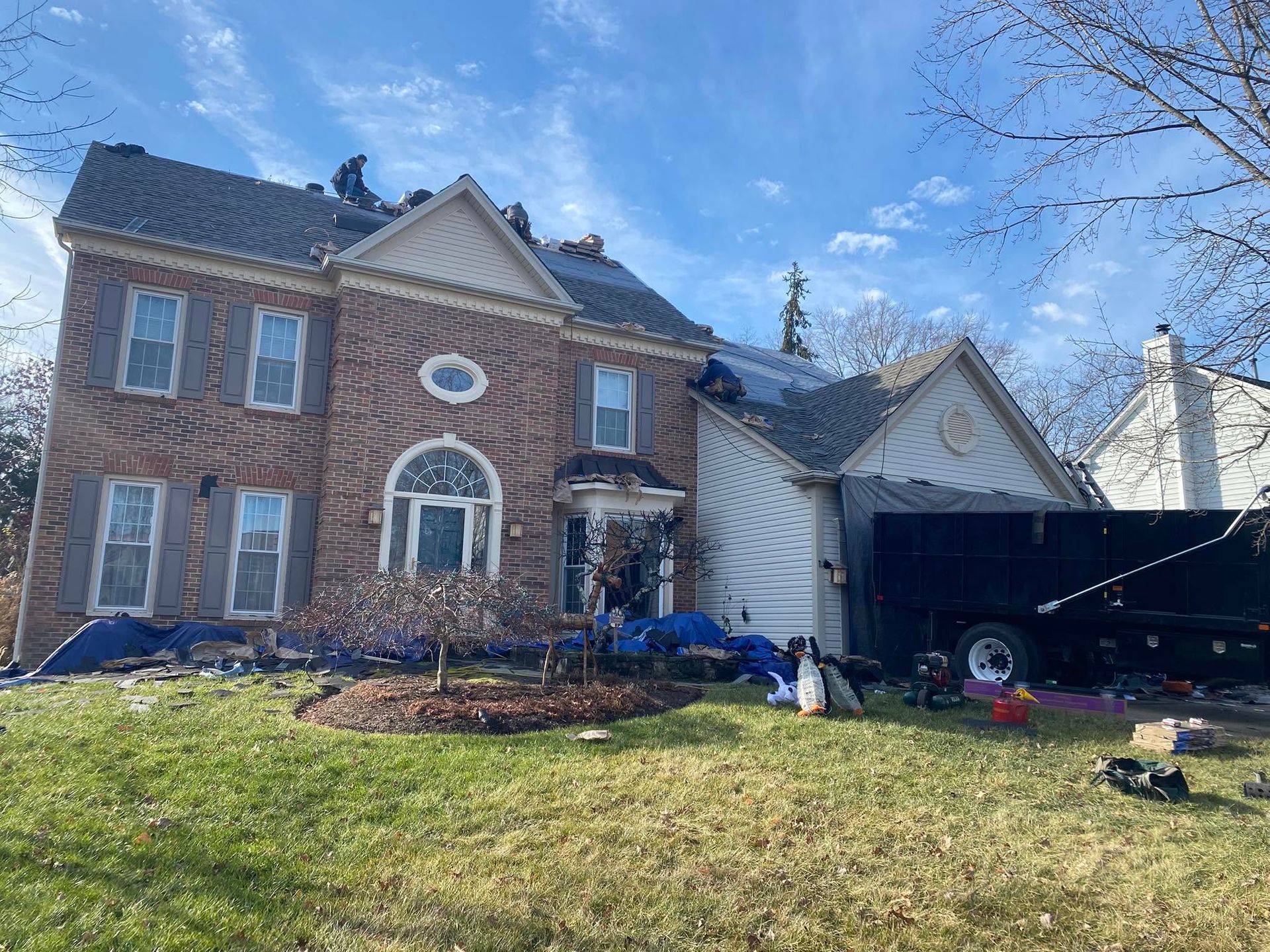 Brick two-story house with blue tarp damage on the right side and debris in the front yard under a clear sky