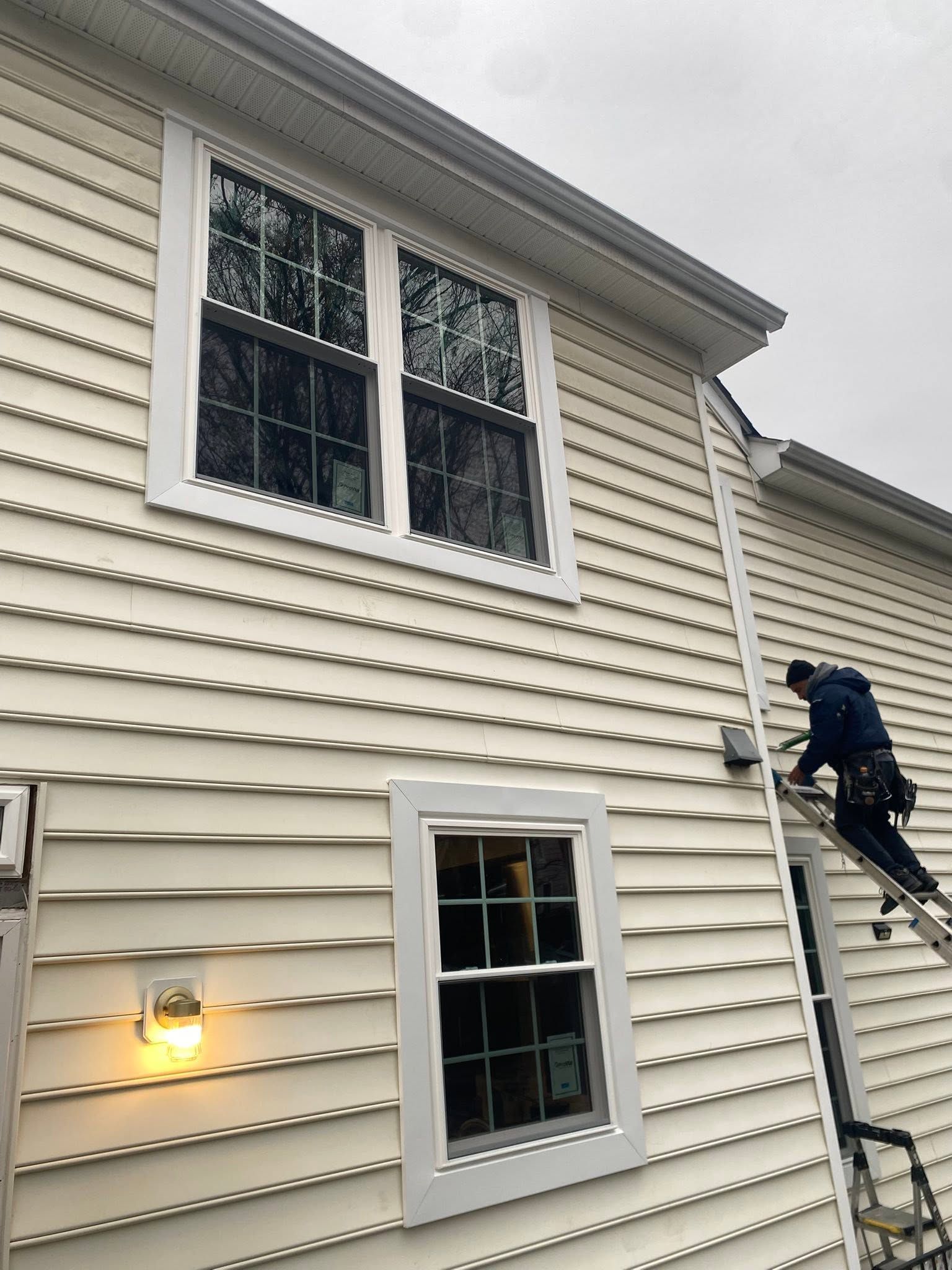 Worker on a ladder painting cream siding on a two-story house near windows.
