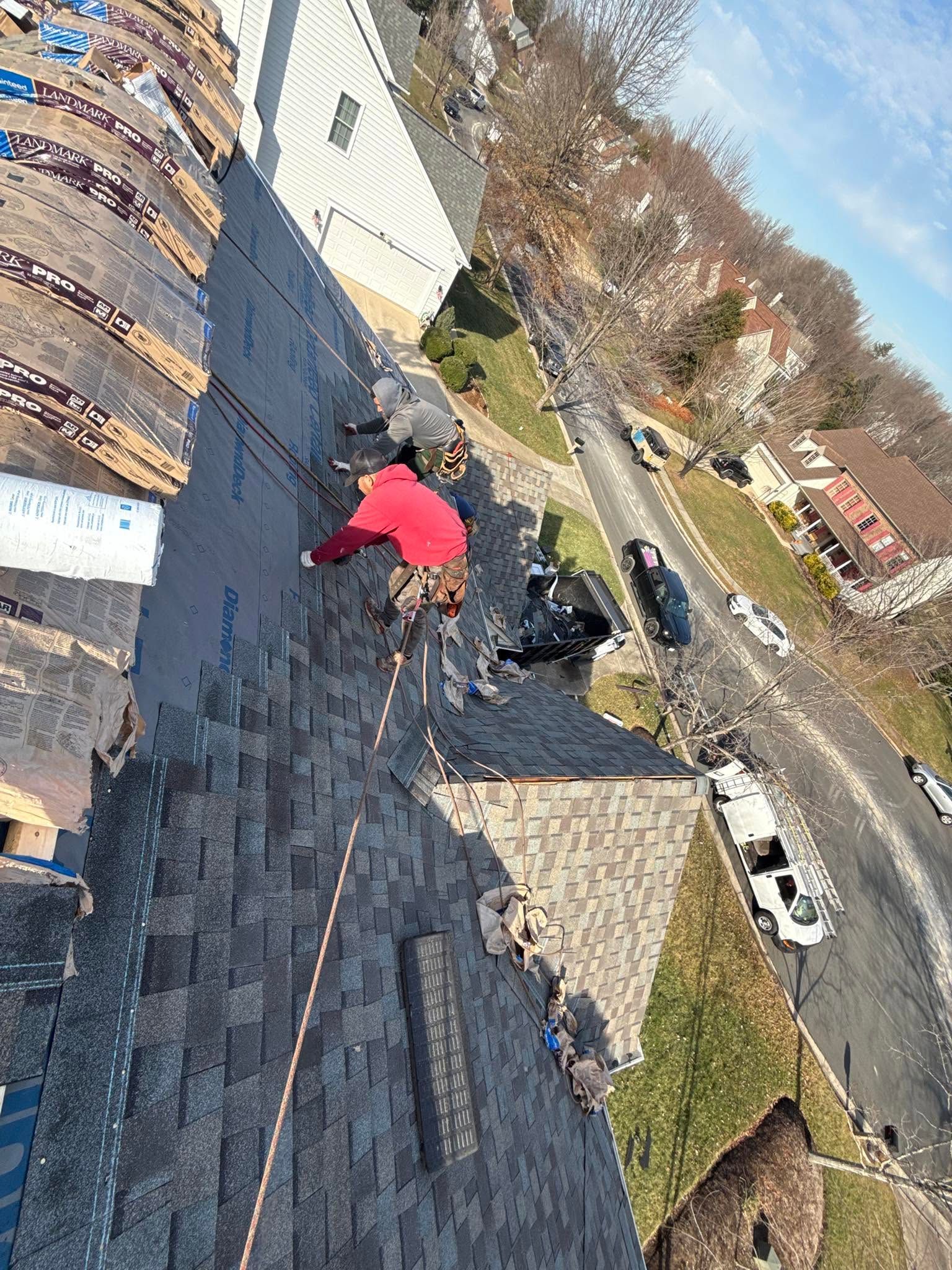 Roof worker in red jacket repairs shingles on a steep rooftop beside a quiet residential street.