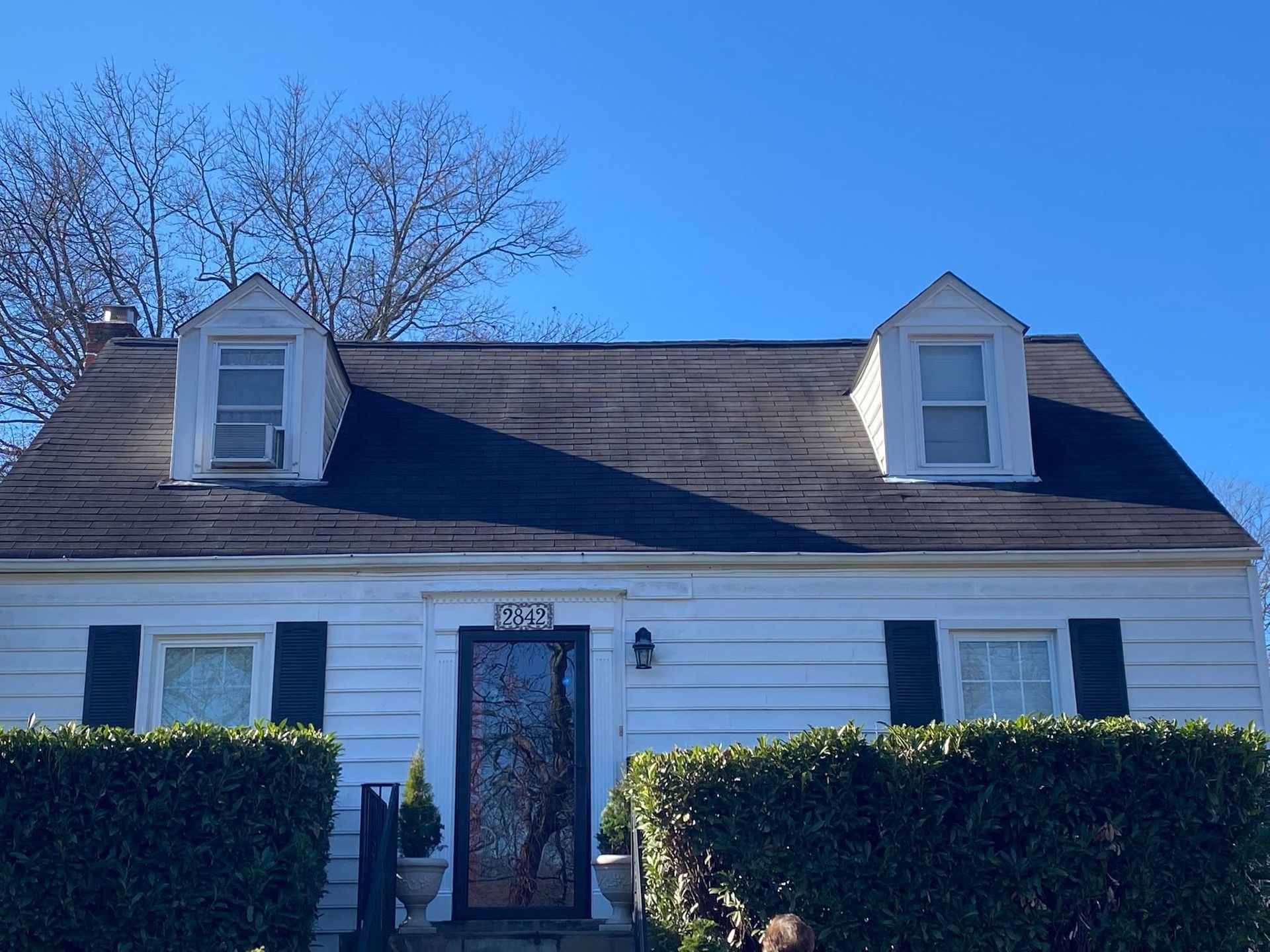 White house with dark roof, two dormer windows, and trimmed hedges under a clear blue sky.