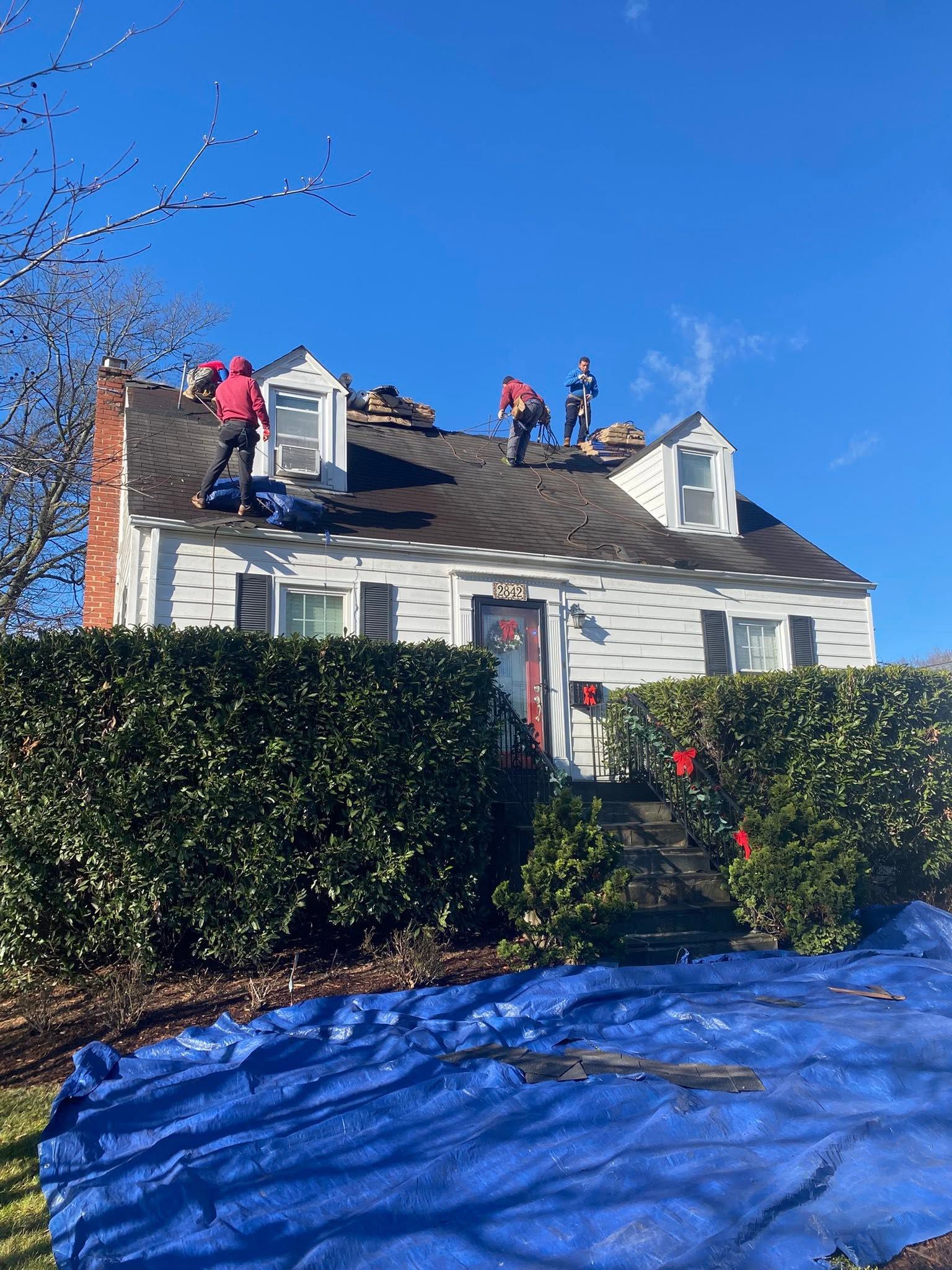 Workers repairing a white house roof with blue tarp and hedges in front, under a clear blue sky