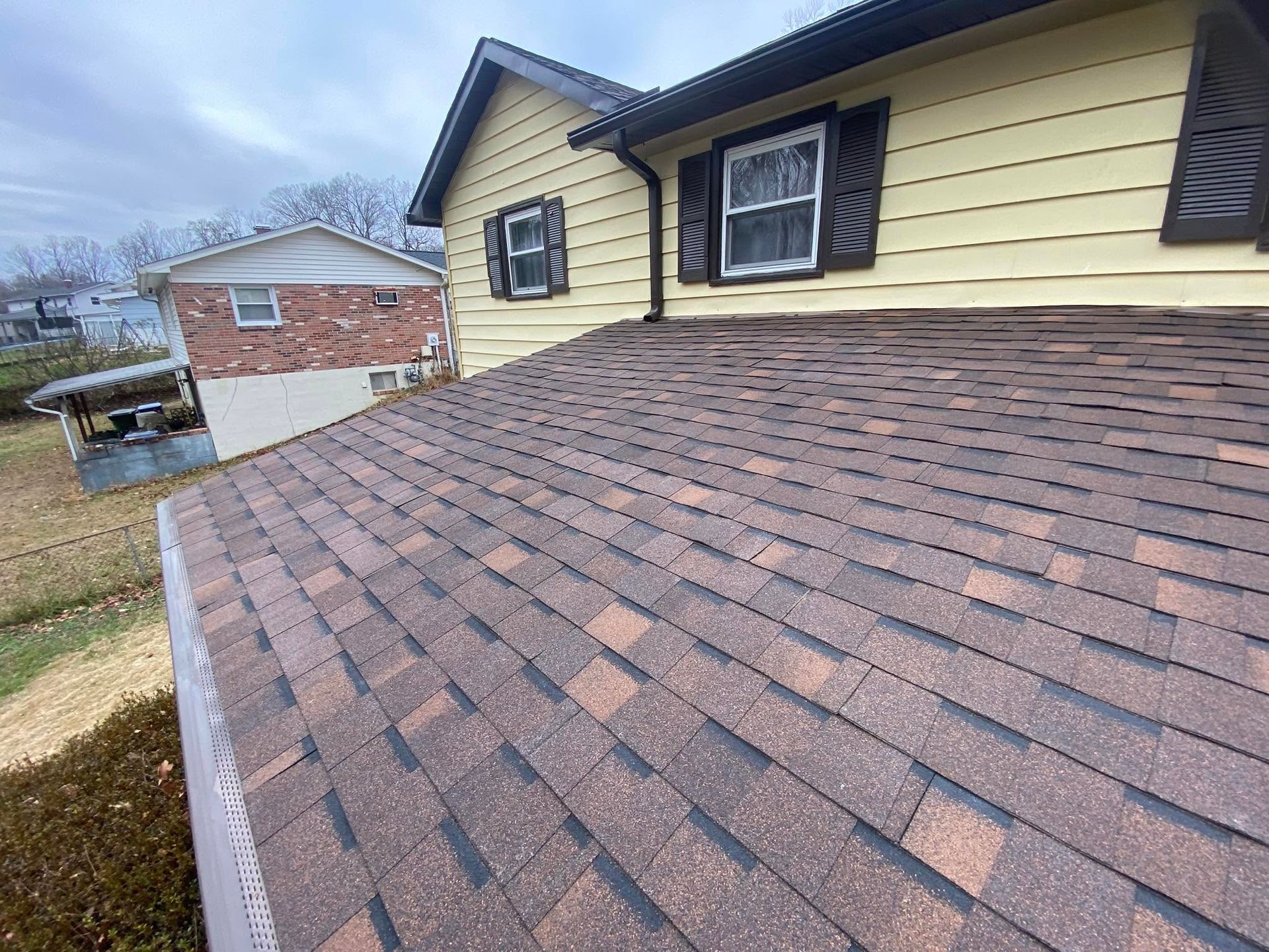New brown shingle roof on a yellow house with dormer windows and a brick garage in the background