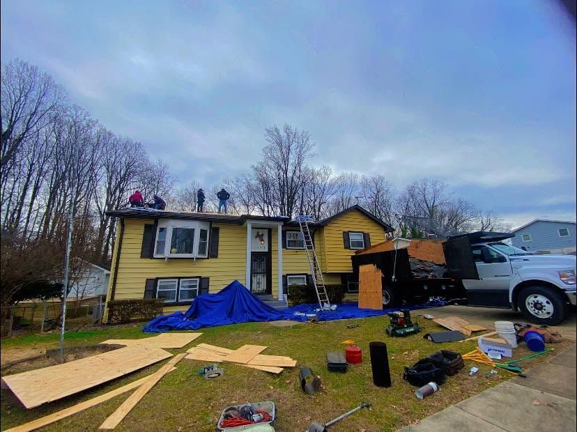 Damaged yellow house with roof work, blue tarp, tools, and a white truck in a yard under a cloudy sky