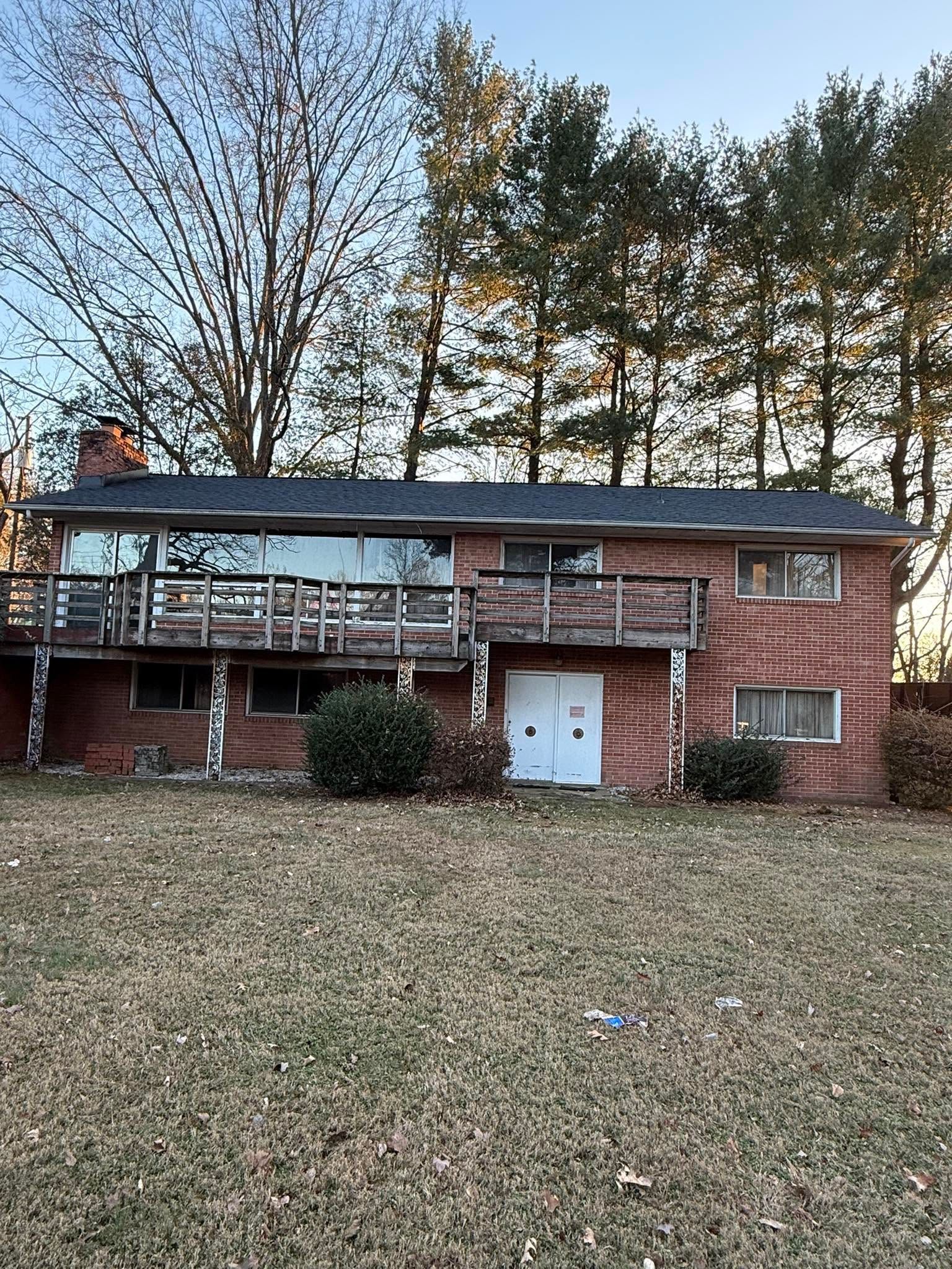 Two-story brick house with deck and white doors, set behind a grassy yard and tall trees