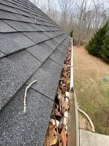 Roof gutter packed with leaves beside a shingled house, viewed along the roofline.