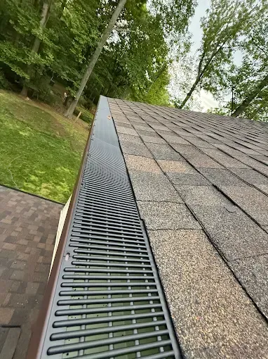 Roof gutter with metal leaf guard along a shingled roof, beside trees and grass.