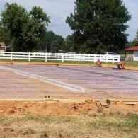 Empty outdoor riding arena with two horses, white fence, and trees under a cloudy sky