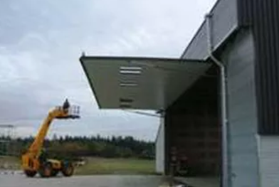 Yellow boom lift raising a worker beside a gray industrial building with an open awning