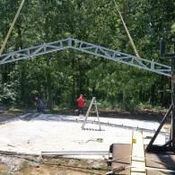 Partially built metal truss arch over a concrete slab with workers and trees in the background