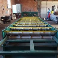 Industrial workshop with a row of metal frames and rollers, with two workers near a car and brick wall.