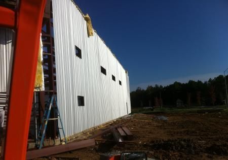 White metal-sided building under construction beside a red frame, with a ladder and dirt site in sunlight.