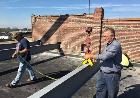 Two workers installing a rooftop beam beside a brick wall on a sunny day