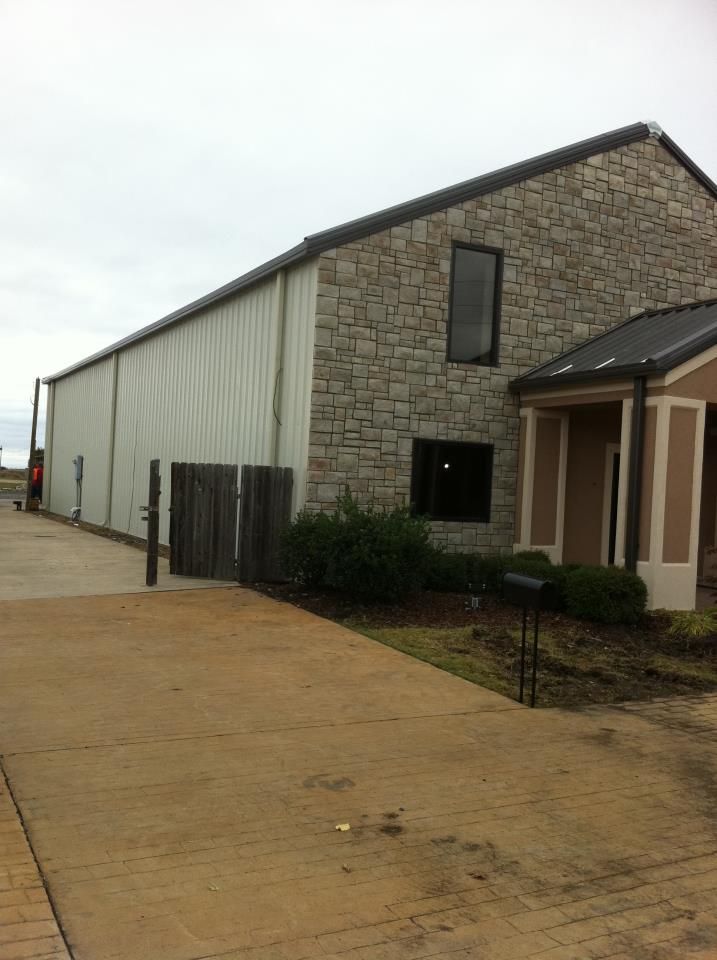 Stone-and-metal industrial building with a driveway and small front shrubs under an overcast sky