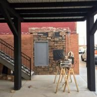 Outdoor work area with a brick wall, mounted window, stacked stone, and a stand holding tools under a covered patio