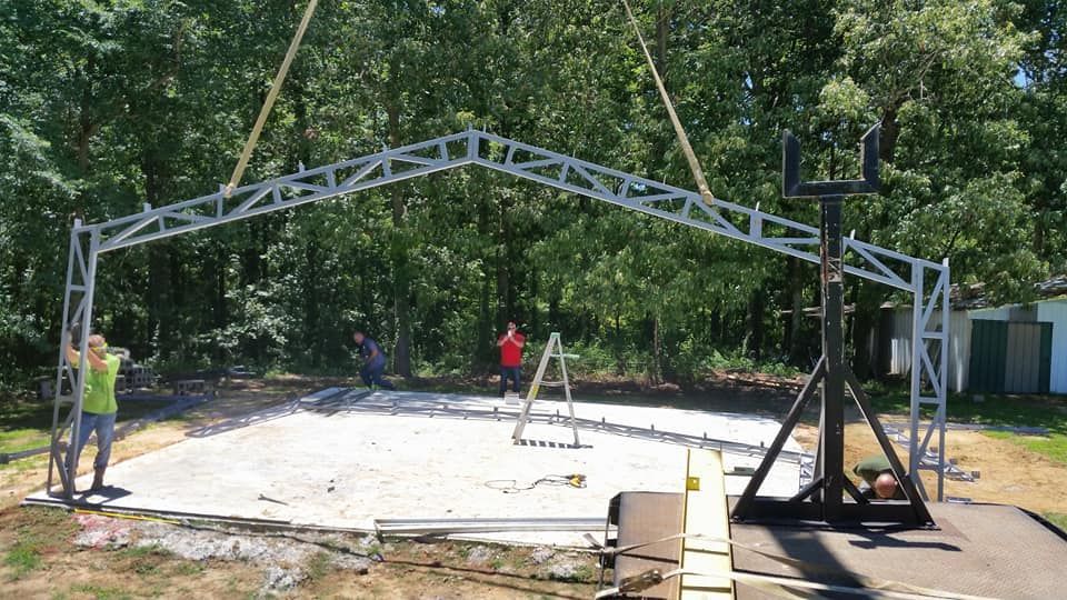 Outdoor construction site with a large metal arch frame being assembled on a concrete pad by workers and equipment