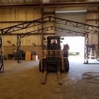 Forklift inside a warehouse, with a person standing beside it and an unfinished metal frame overhead.