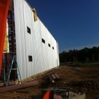 White metal-sided building under construction beside a dirt lot, with ladder and blue sky.