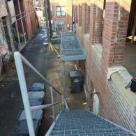 Narrow outdoor walkway with brick walls, metal grates, and construction materials along the side of a building.