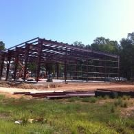Steel frame of a building under construction on a cleared site, with grass and trees nearby.