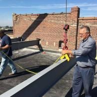 Two workers lifting a long gray beam on a rooftop using a red crane hoist