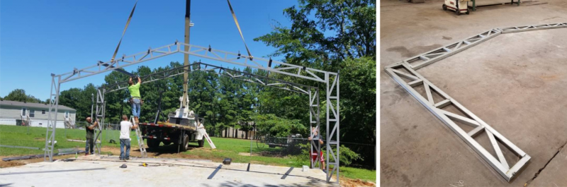 Crane lifting a triangular metal truss outdoors, with workers and a construction site in the background.