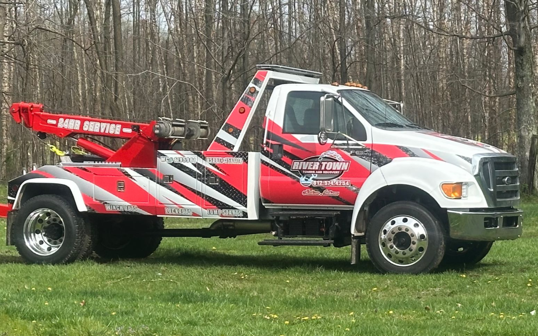 Red and white tow truck parked on grass beside a wooded area