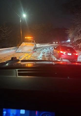 Nighttime road scene with a tow truck and a red car in snowy conditions, viewed from inside a car.