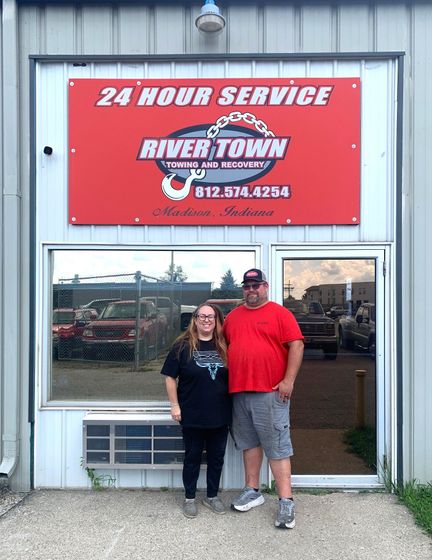 Two people standing outside a shop with a red “24 Hour Service” River Town Towing sign above the door