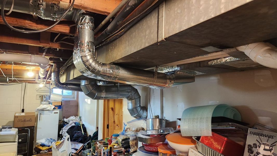 Basement cluttered with exposed ductwork, pipes, storage items, and a wooden door under low ceiling beams.