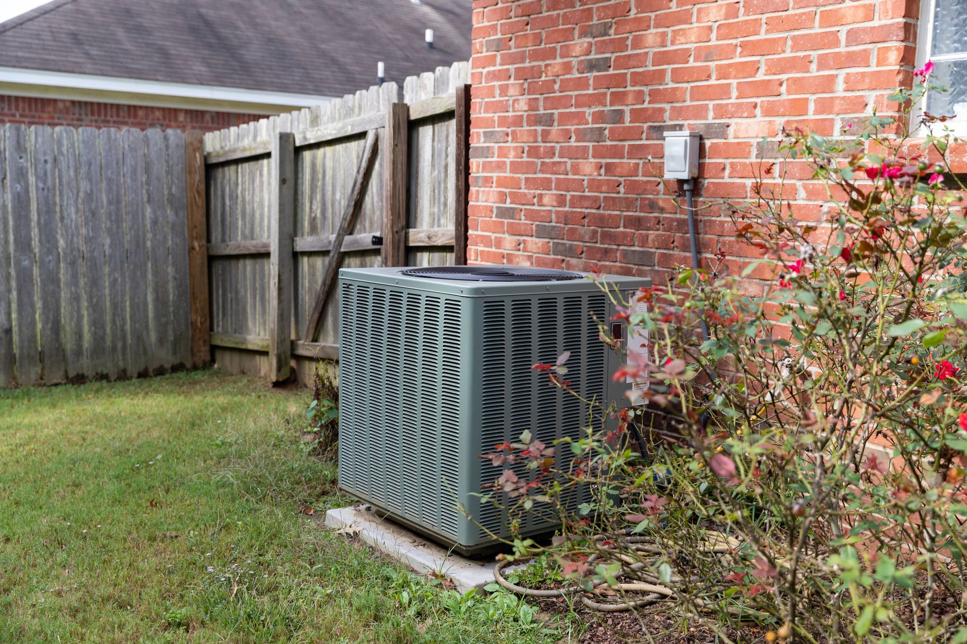 Outdoor AC unit beside a brick house, fence, and shrubs in a backyard