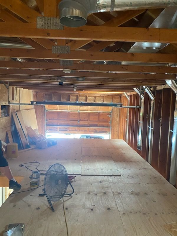 Unfinished attic with exposed beams, plywood floor, tools, and bright sunlight from the far end.
