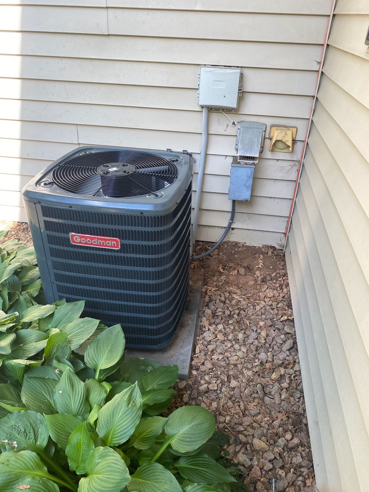 Outdoor air conditioner unit beside house siding with utility boxes and rocks, next to green plants