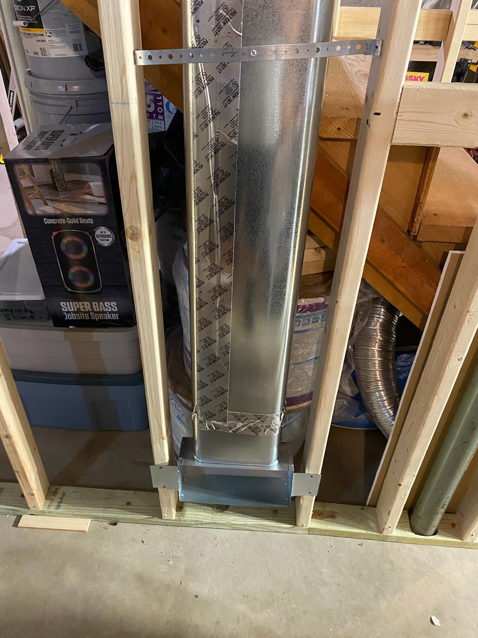 Vertical metal duct/vent framed by wood studs in an unfinished wall, with a concrete floor below.