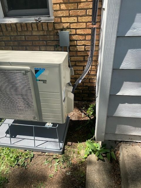Outdoor HVAC unit beside brick wall and gray siding, with electrical conduit and plants nearby.