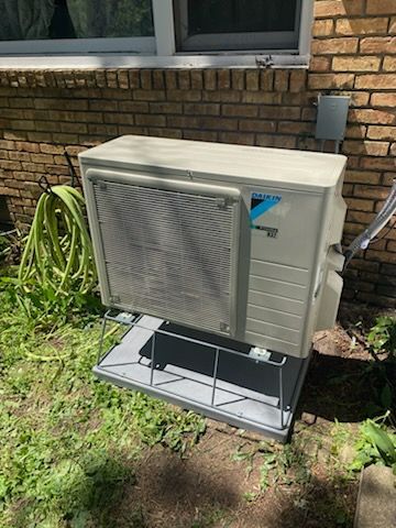 Outdoor air-conditioning unit beside a brick wall and window, on a metal stand in a grassy yard.