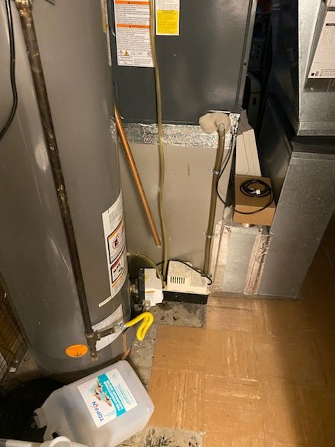 Water heater in a cramped utility closet with pipes, a hose, and a cardboard box on the floor.