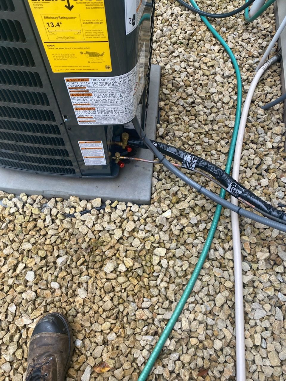 Outdoor HVAC unit on gravel beside a green hose and black cables, with a boot visible in the corner