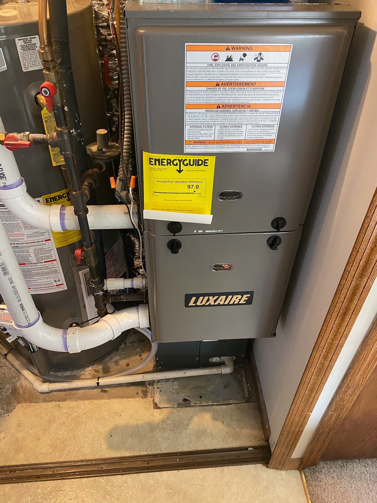Gray furnace in a utility closet with pipes and warning labels, near a doorway and wood wall.