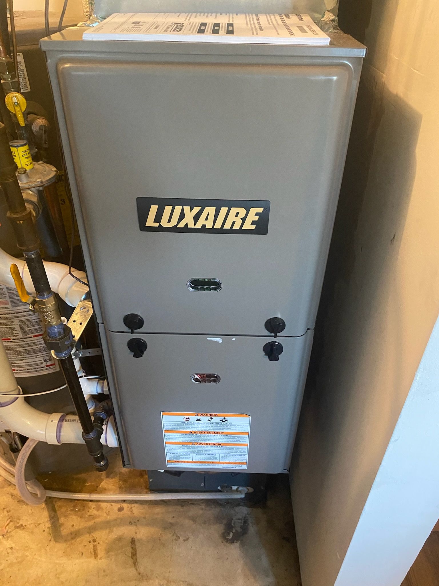 Gray Luxaire HVAC furnace in a narrow utility closet with pipes and wiring visible.