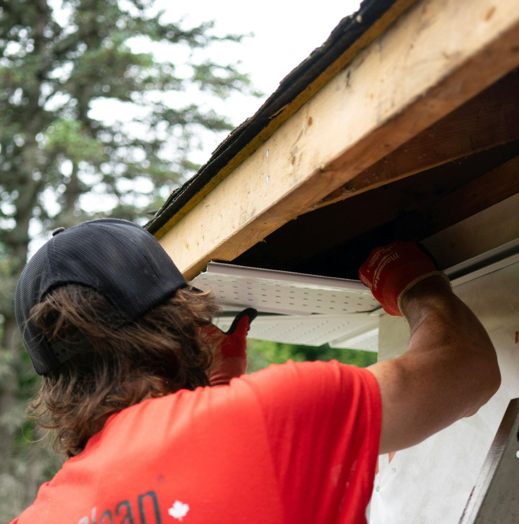 Person in red shirt and cap working under a roof eave with a tool.