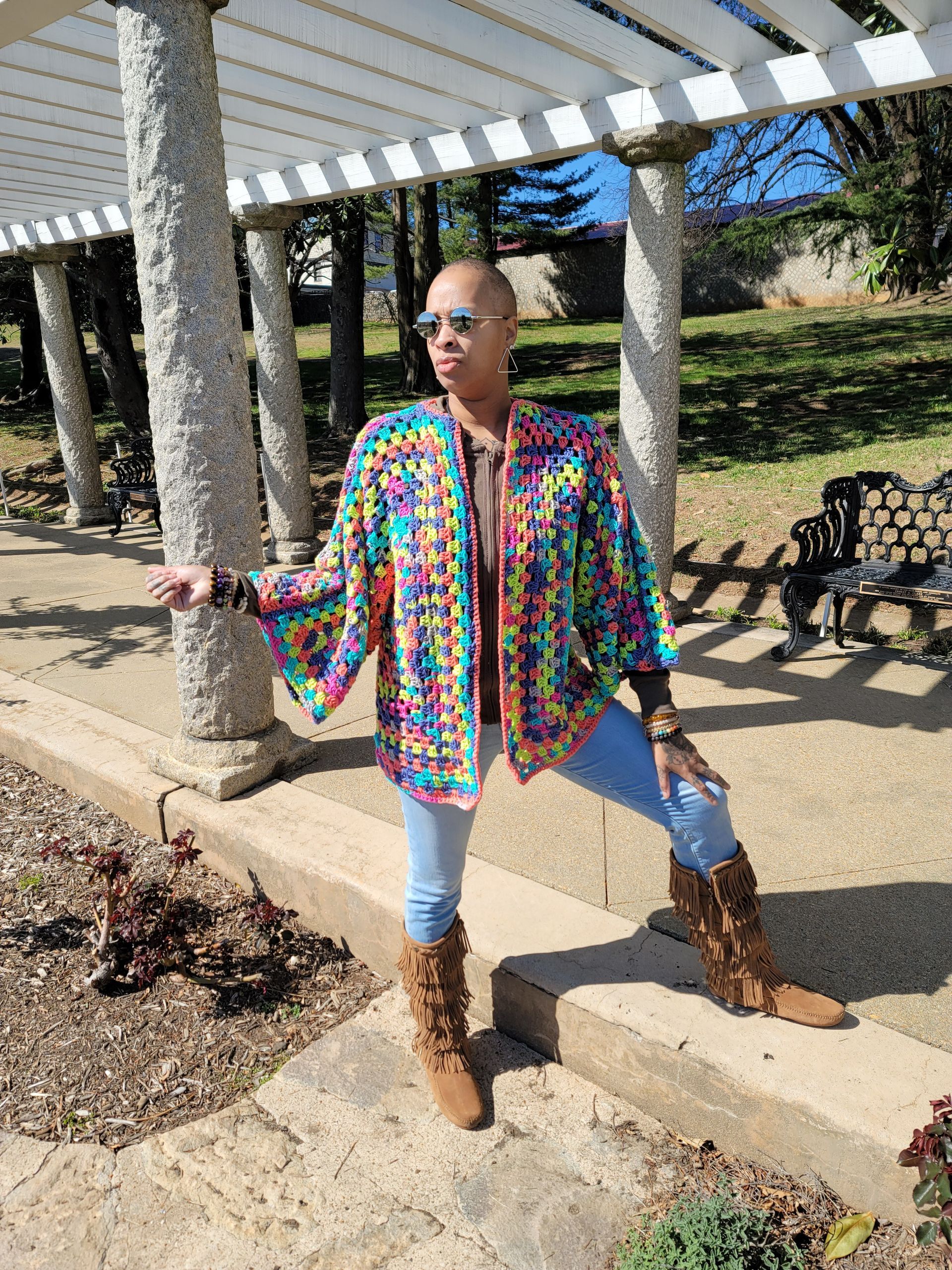 A woman wearing a colorful jacket and boots is standing under a pergola in a park.