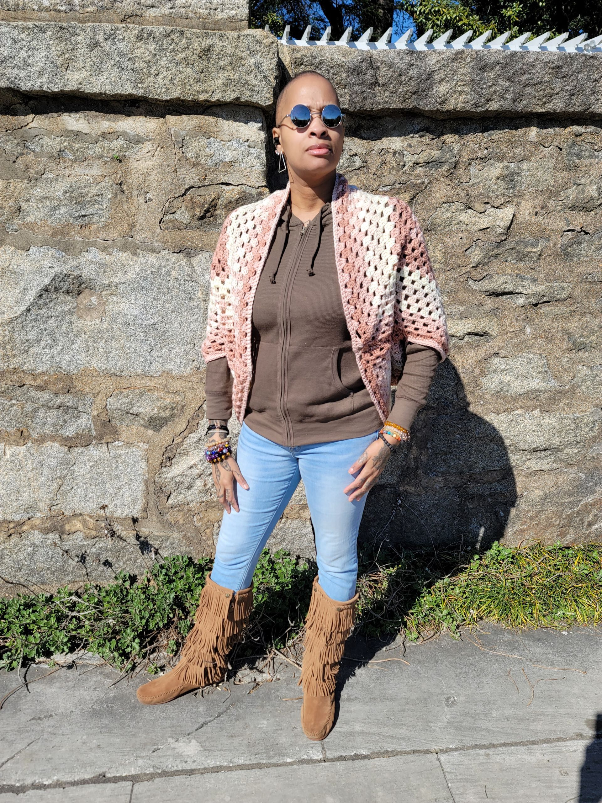 A woman wearing sunglasses and a crocheted jacket is standing in front of a stone wall.