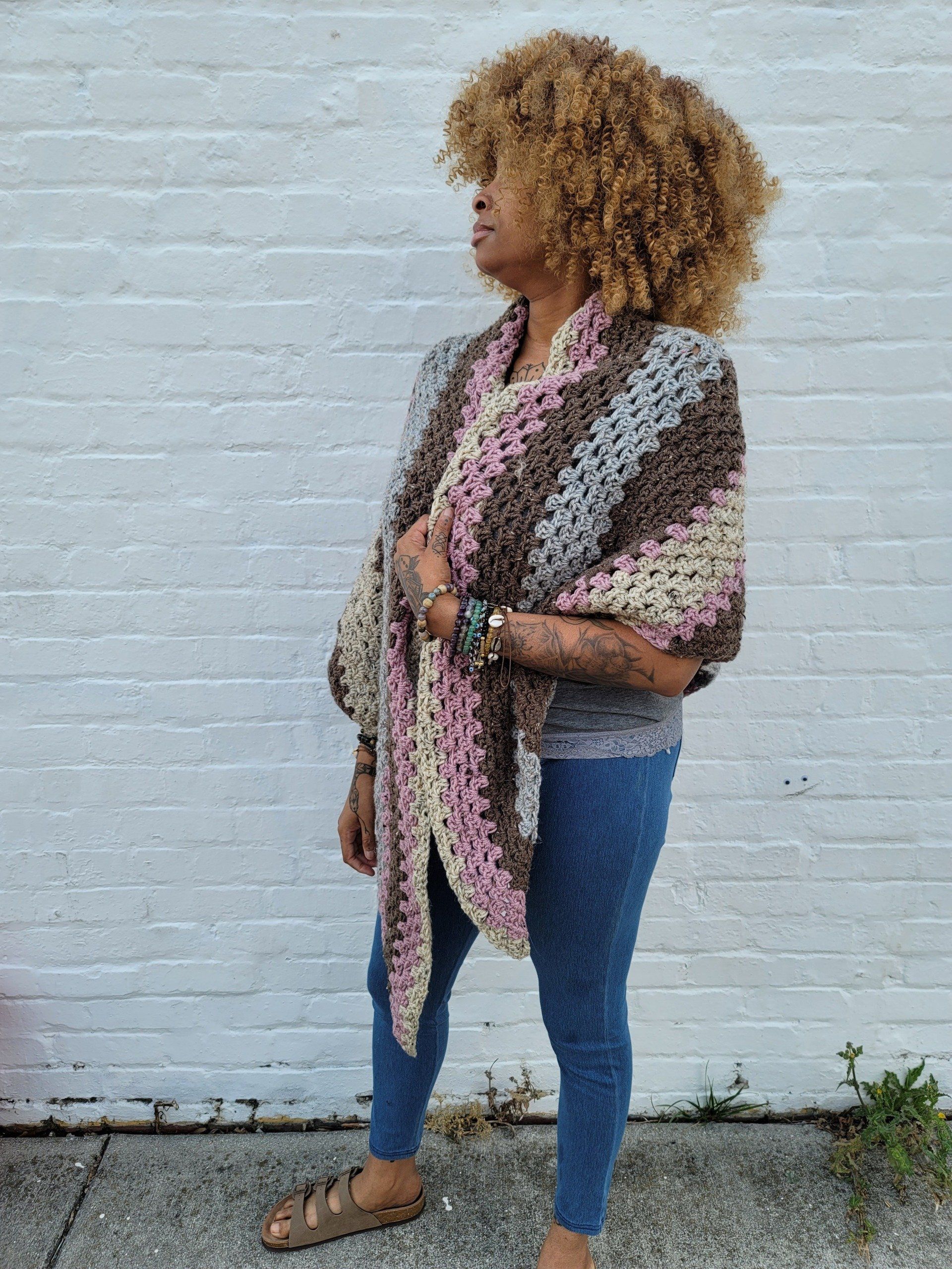 A woman wearing a crocheted shawl is standing in front of a white brick wall.