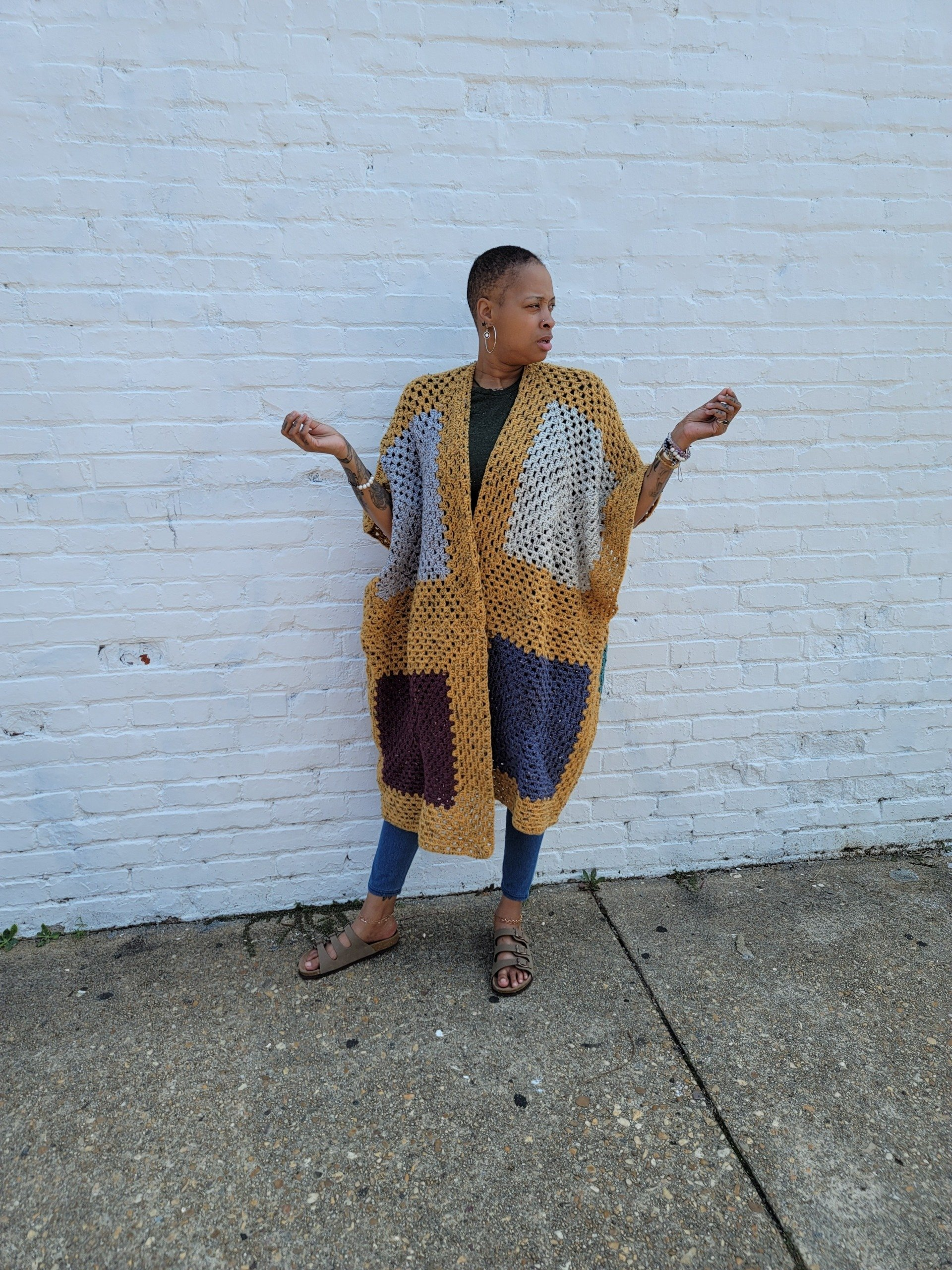 A woman is standing in front of a white brick wall wearing a crocheted cardigan.