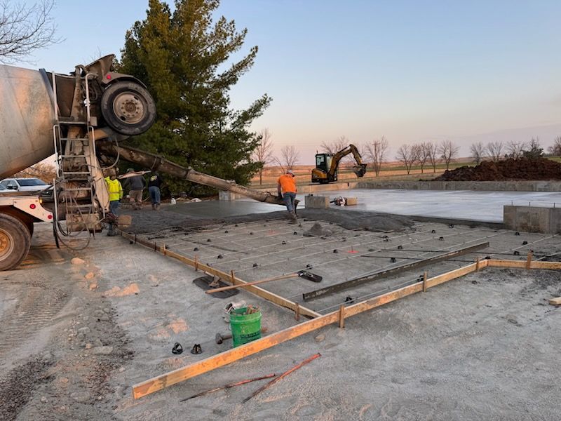 Concrete slab being poured at a construction site with workers, a mixer truck, and an excavator.