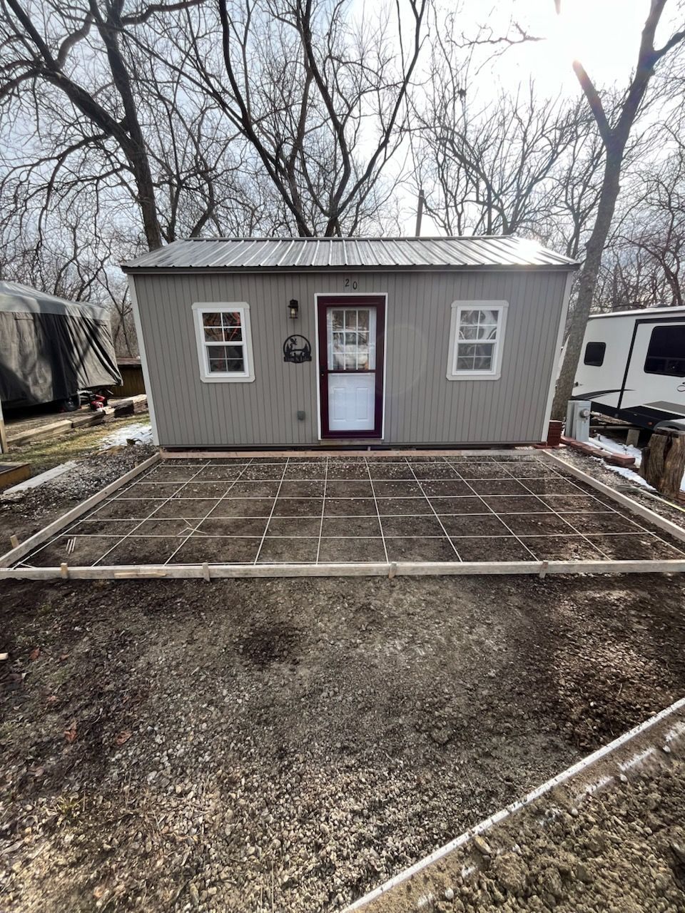 Small gray shed with white trim and a central door, set behind a gravel yard and wooden frame in winter.