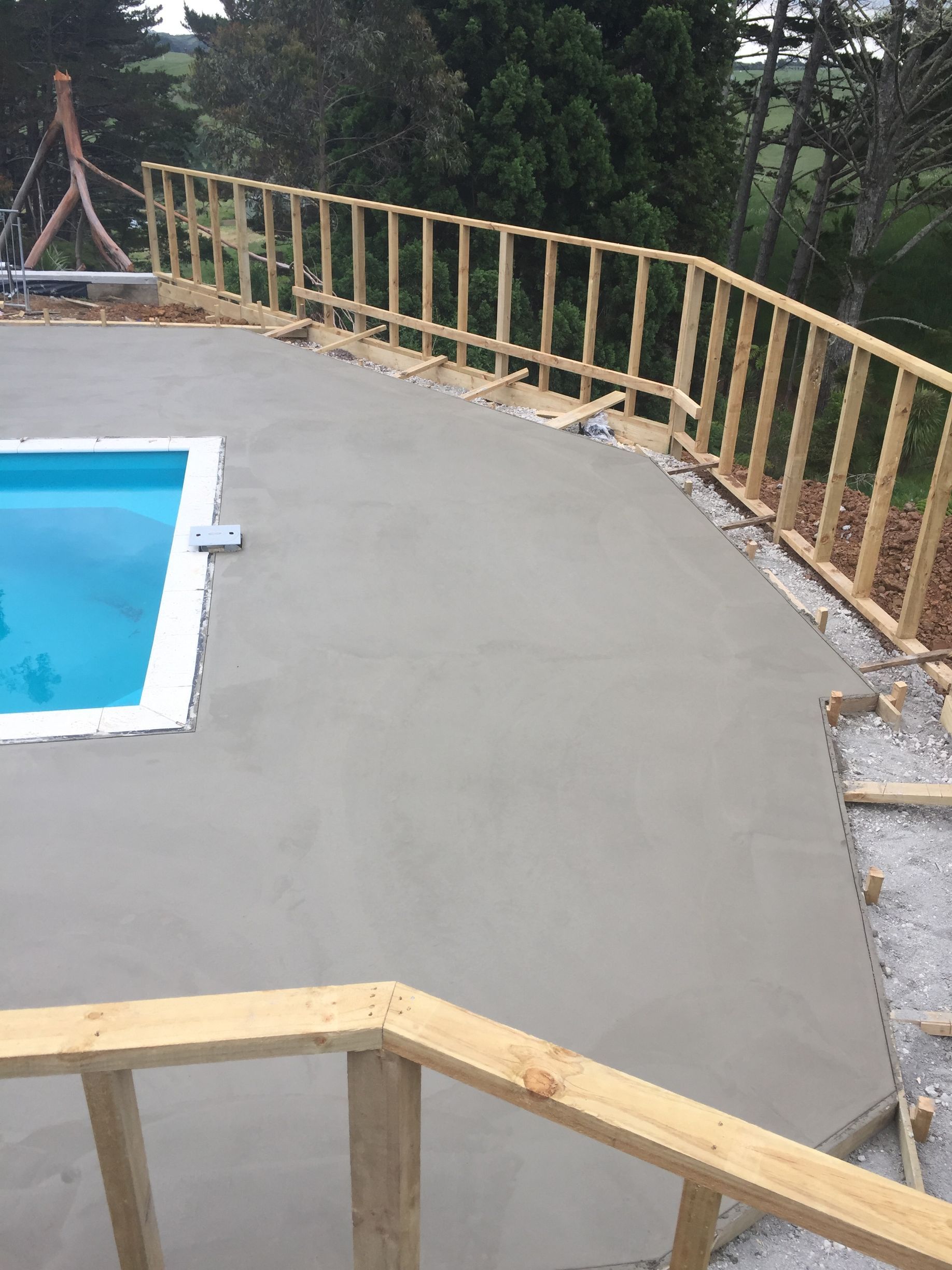Concrete pool deck with blue swimming pool and wooden railing overlooking a wooded area